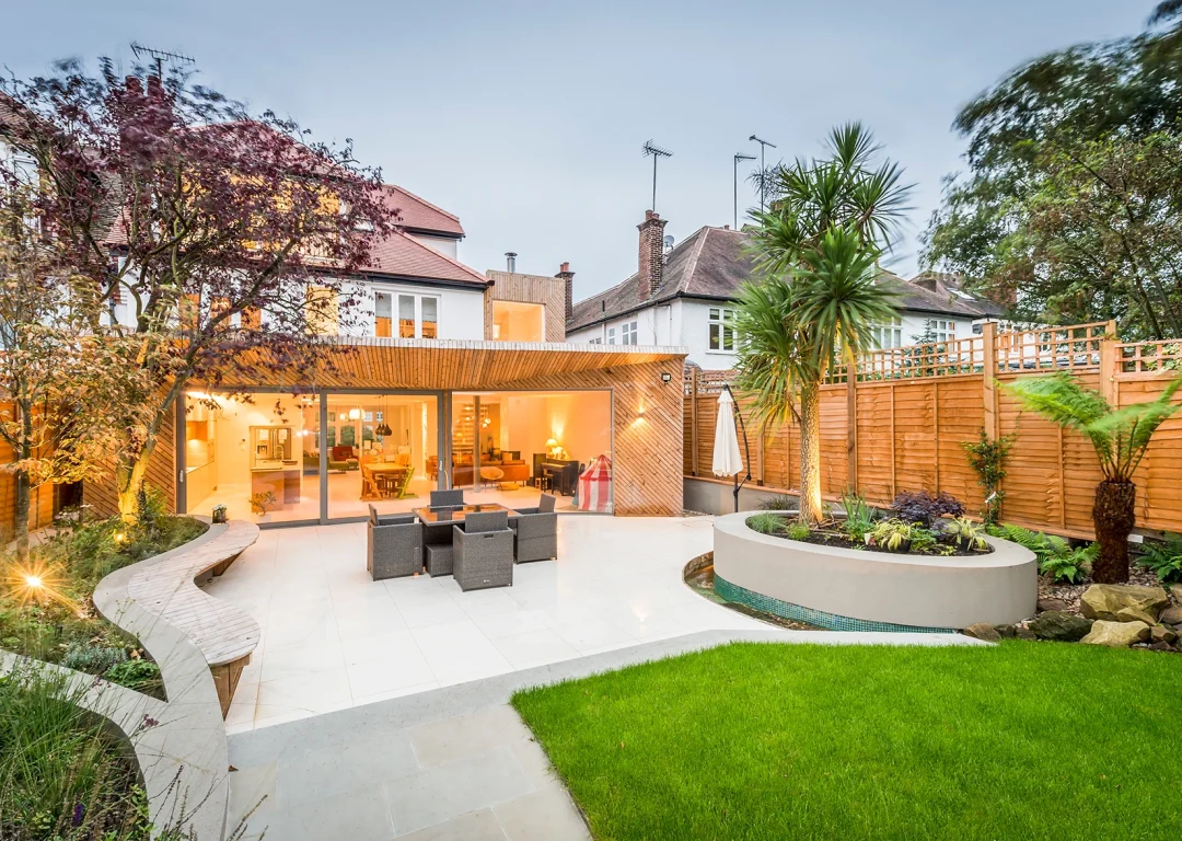 Modern landscaped garden featuring a curved patio with outdoor dining furniture, green lawn, tropical plants, and raised flower beds.