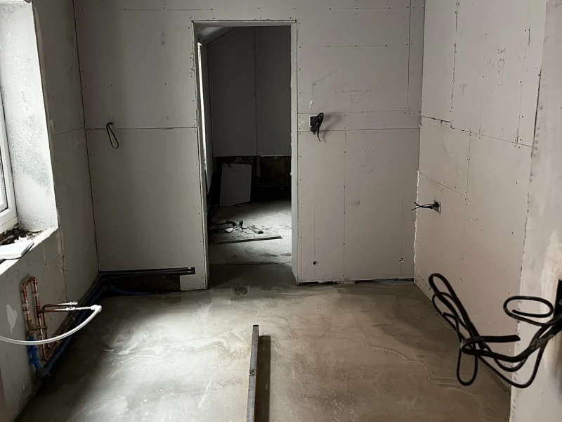 A room renovation featuring a concrete floor and a single door on one wall with electrical wires hanging out of the ceiling.